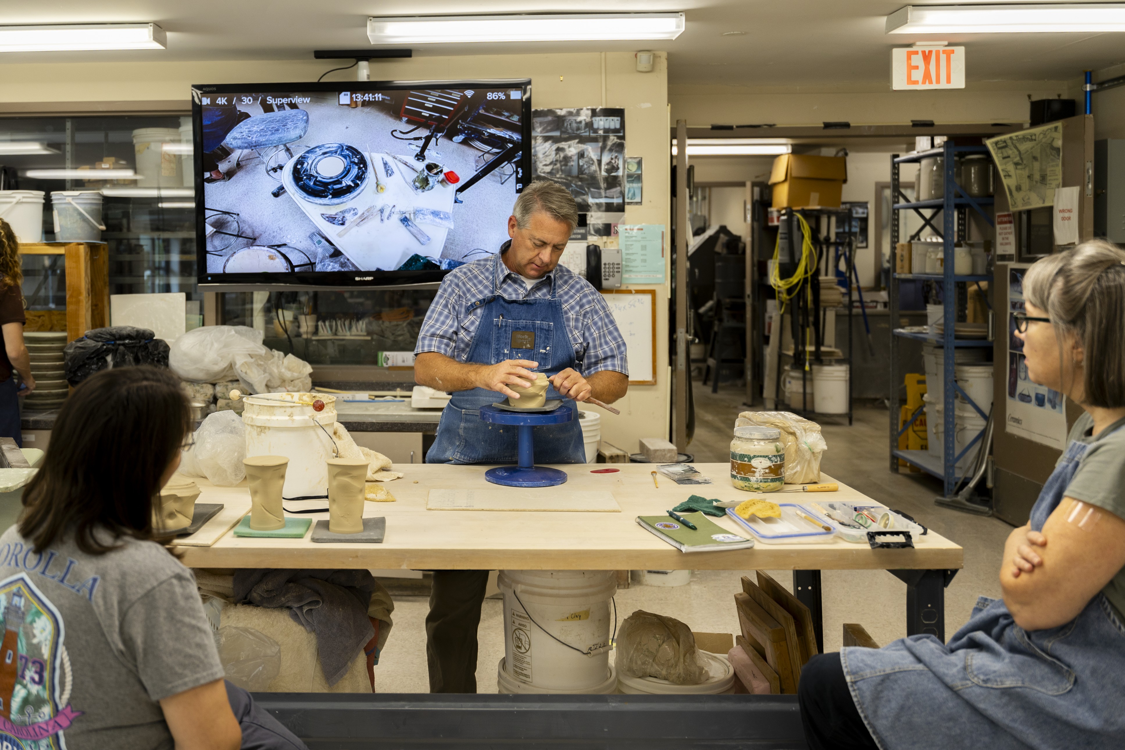A pottery workshop where an instructor shapes a clay vessel on a small wheel at a worktable, while two seated participants watch. Shelving with tools and supplies and a screen showing a close-up of the work area are visible in the background.