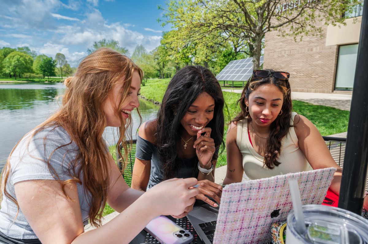Three female students looking at a laptop outside in front of lake huntoon