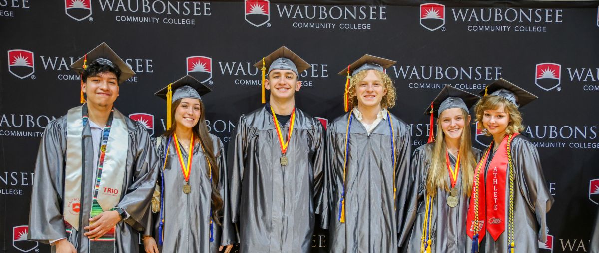 2026 grad hero image six students posing in cap and gown in front of Waubonsee backdrop