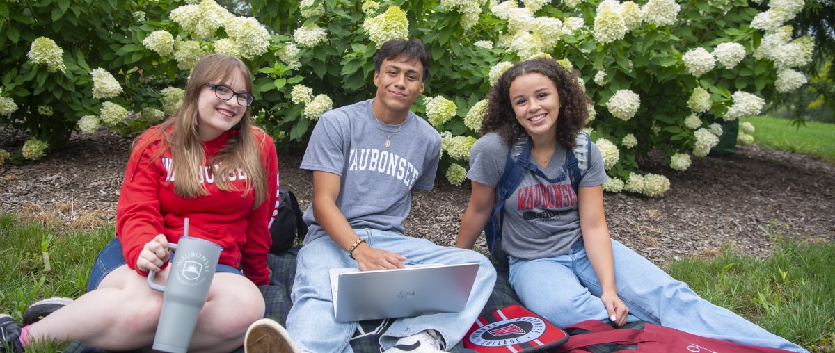 Three students sitting on a blanket outside in front of flowers