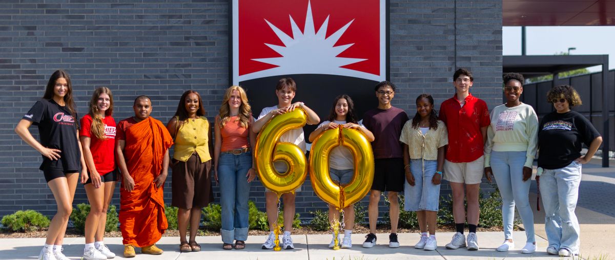 Several students holding balloons in the shape of 60 in front of Waubonsee logo on a building