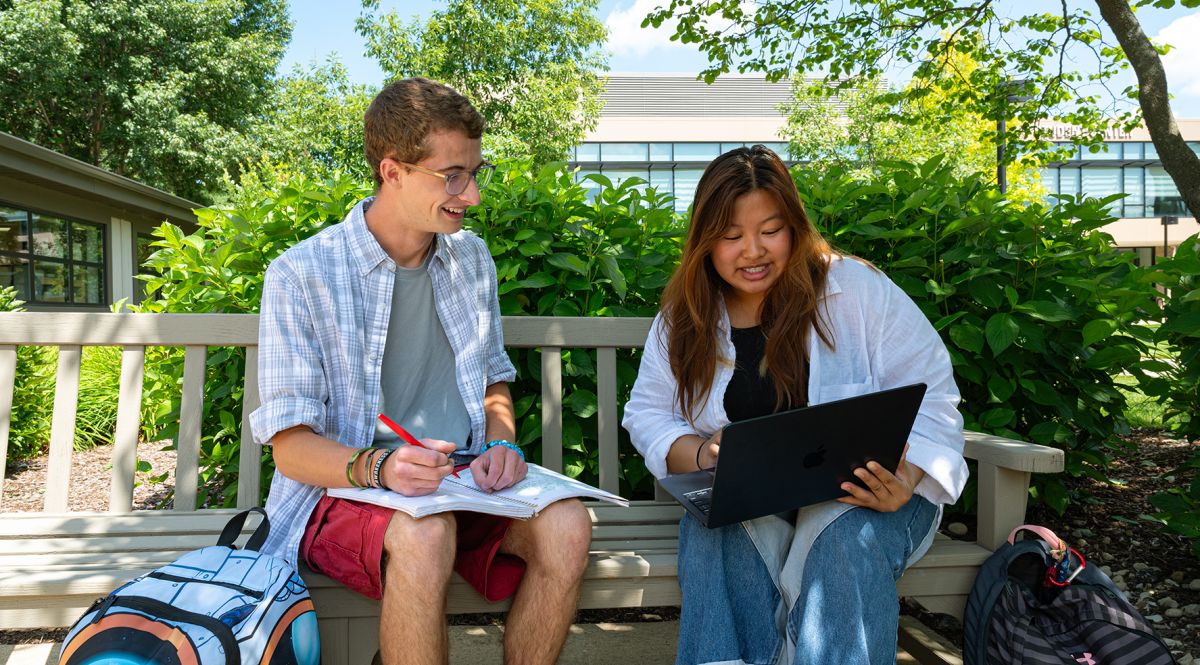 two students sitting outside on bench during summer, one with notepad and the other with a laptop