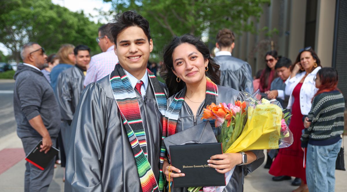 Two graduates wearing caps, gowns, and Hispanic heritage stoles stand outdoors after a graduation ceremony, holding a diploma and flowers.