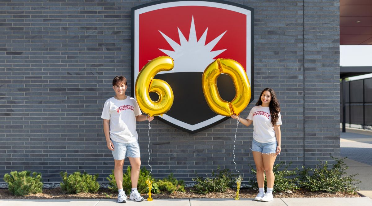 Two students holding balloons in the shape of 60 in front of Waubonsee logo on a building