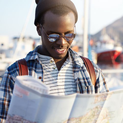 Happy excited dark skinned male presenting traveler wearing stylish clothing holding city-guide in his hands while thinking about where to go