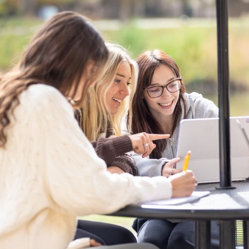 Three female students sitting at table in front of lake Huntoon, looking at the same laptop.